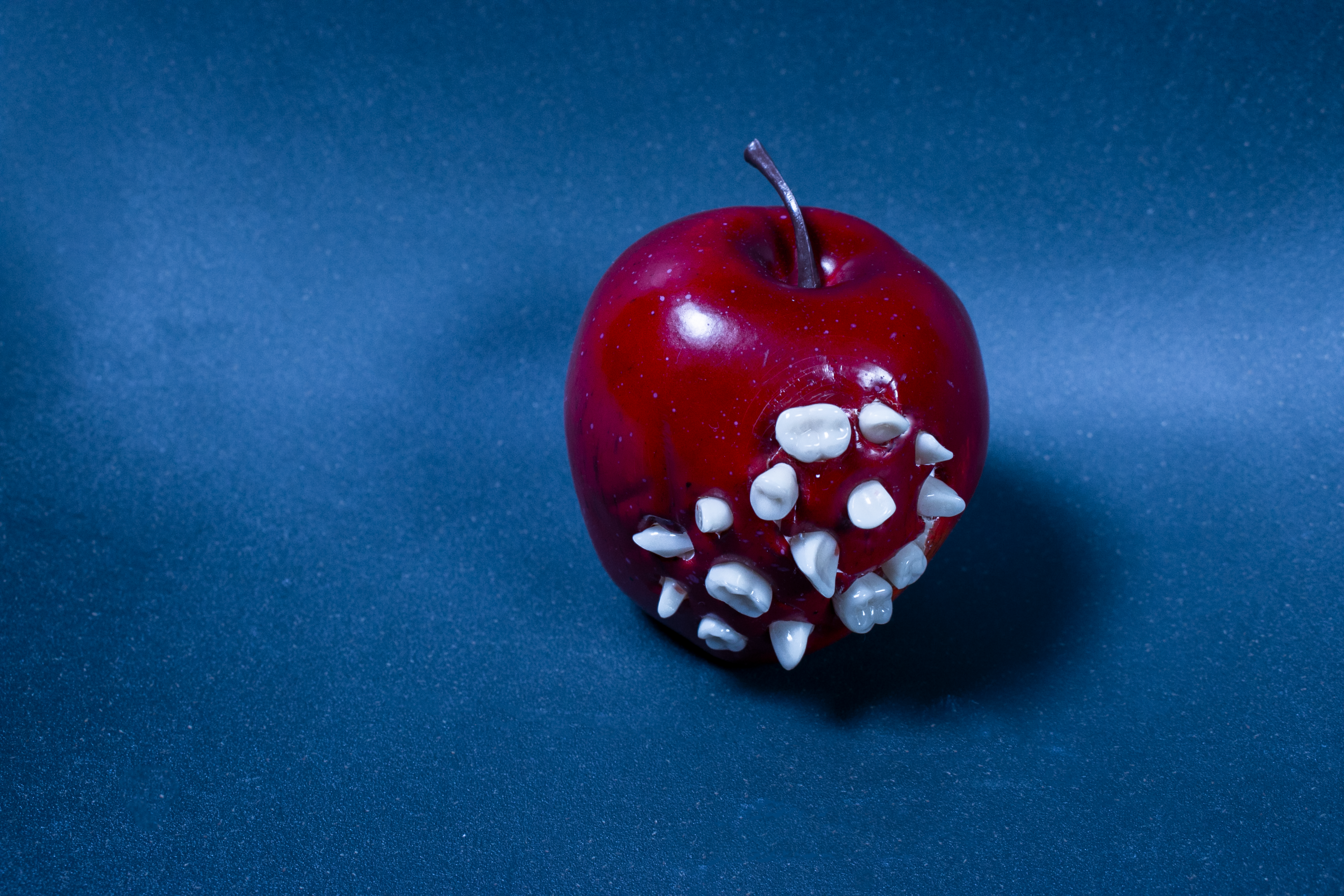 A glossy red apple studded with human teeth on a blue ground